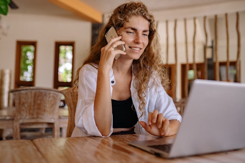 Smiling woman working remotely with a laptop and phone in a cozy setting.