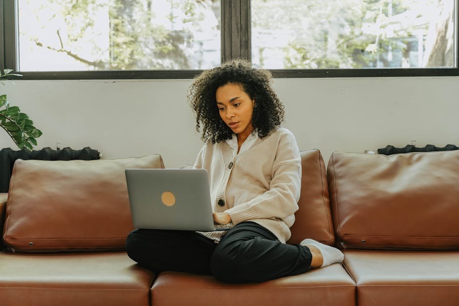 A woman with curly hair sits comfortably on a sofa, working on her laptop indoors.