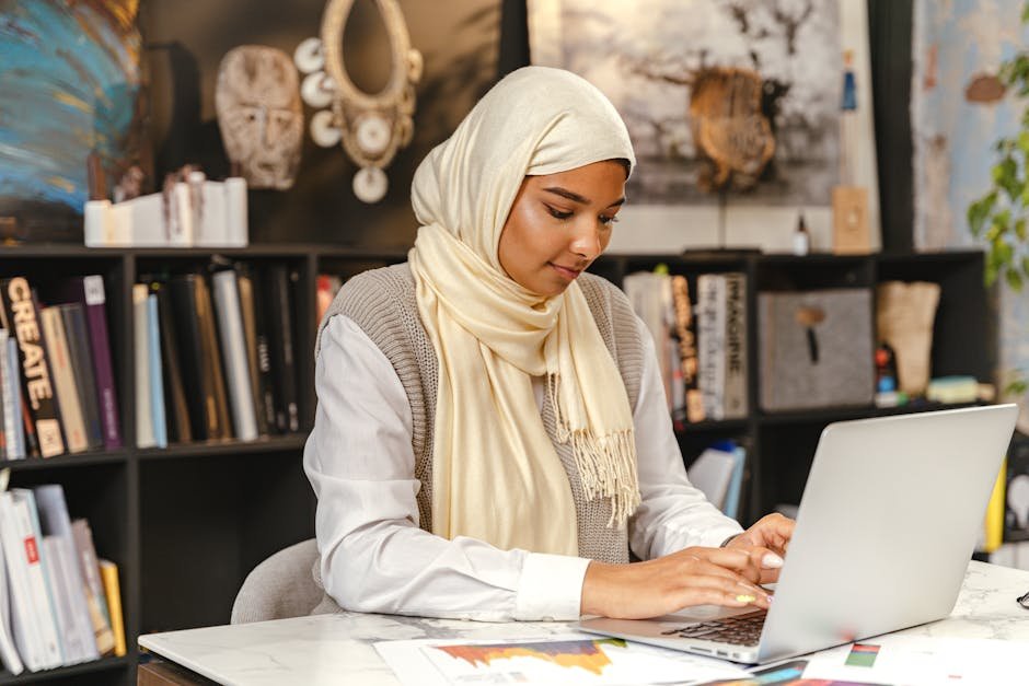 A Muslim woman in a hijab working on a laptop in a contemporary office setting, focused and professional.