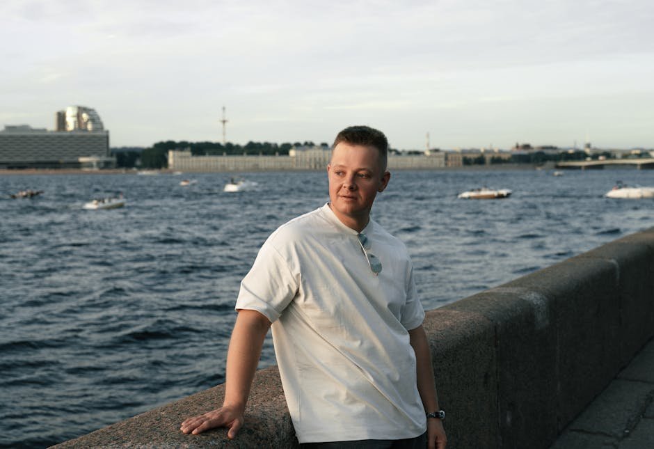Young man in casual attire standing by a river with cityscape in the background
