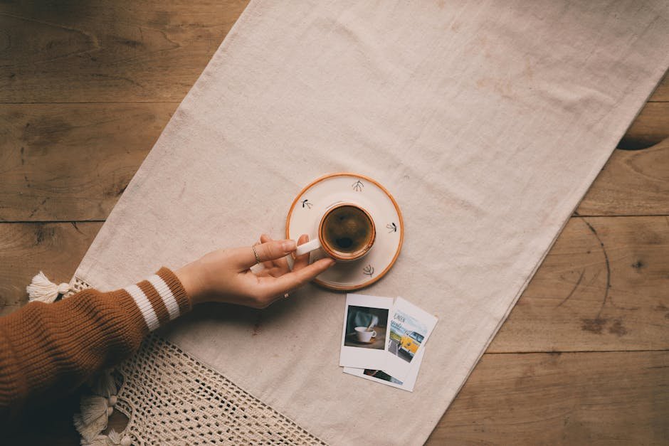 Hand holding coffee cup over linen cloth with polaroids. Cozy and warm ambience.